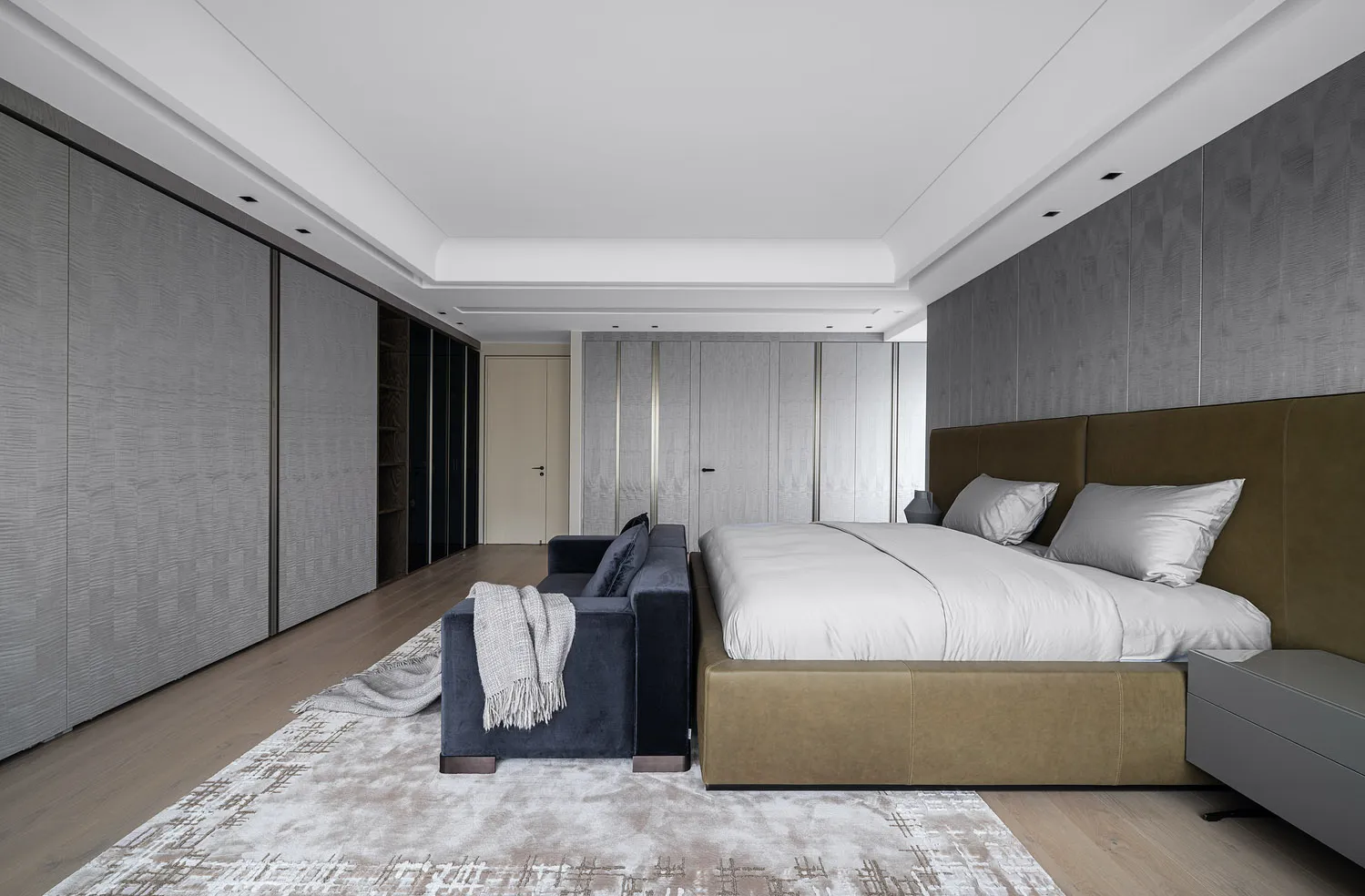 A minimalist bedroom featuring a beige leather bed against a gray wall, with a blue velvet sofa and a textured rug.