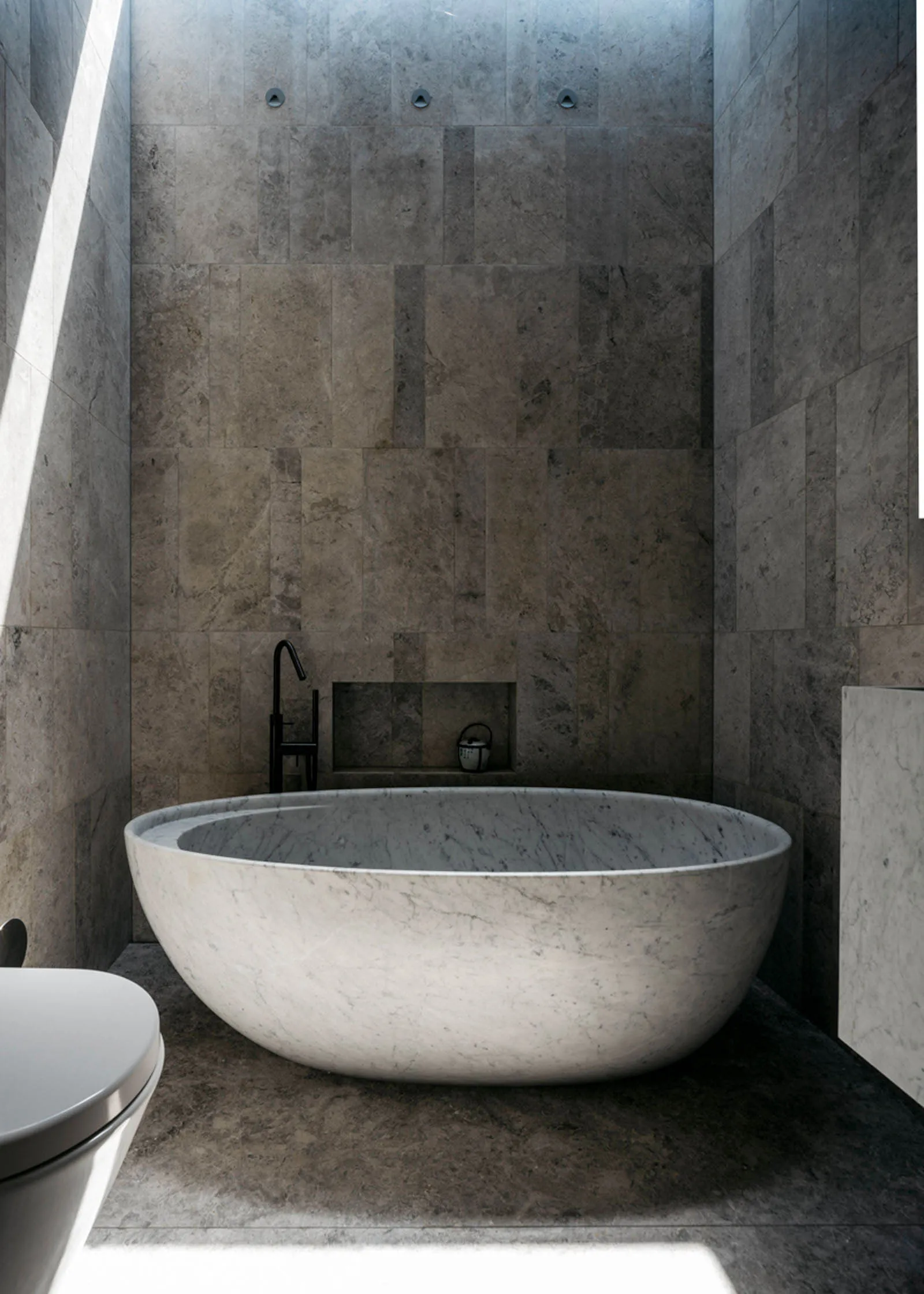 A modern bathroom with a minimalist design, featuring a freestanding marble bathtub and gray marble tiles on the walls.