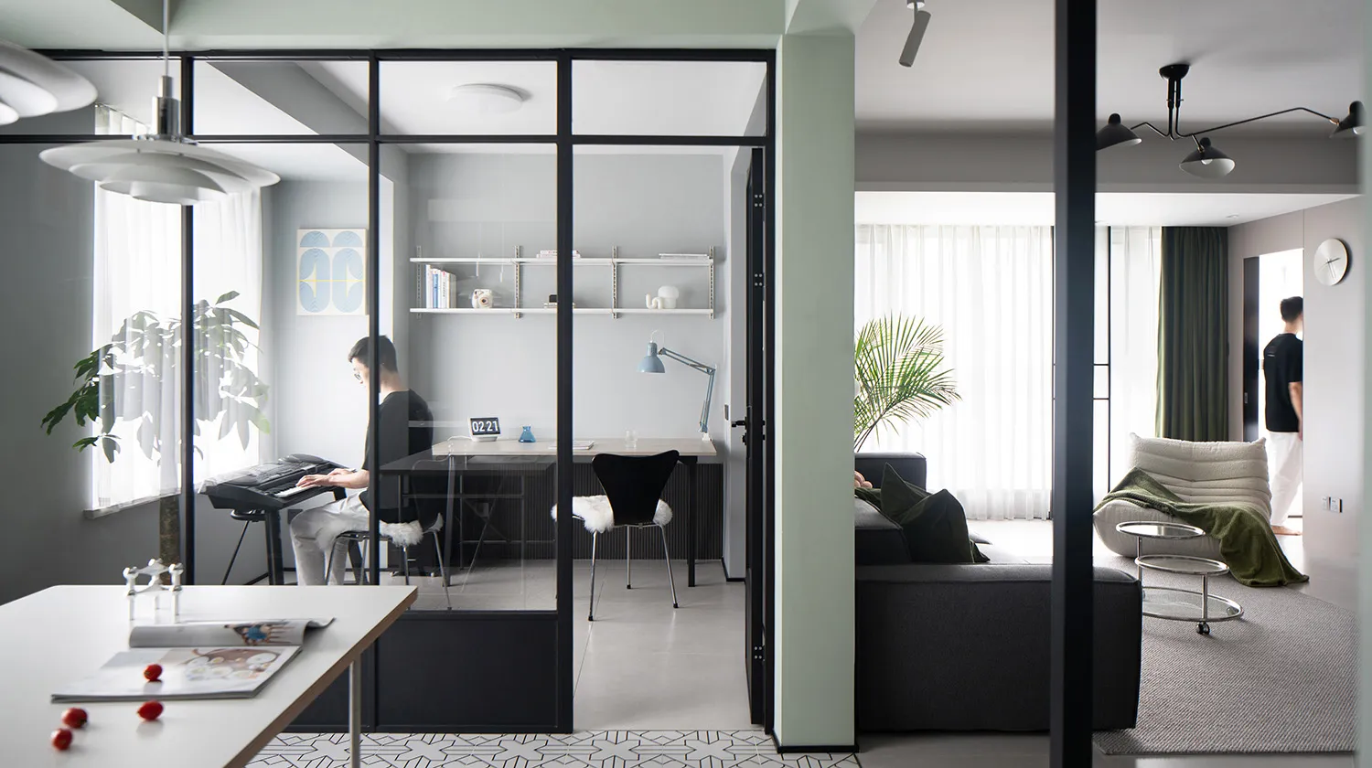 A modern, minimalist apartment office space with a transparent glass wall, black office chair, and a black sofa in the living