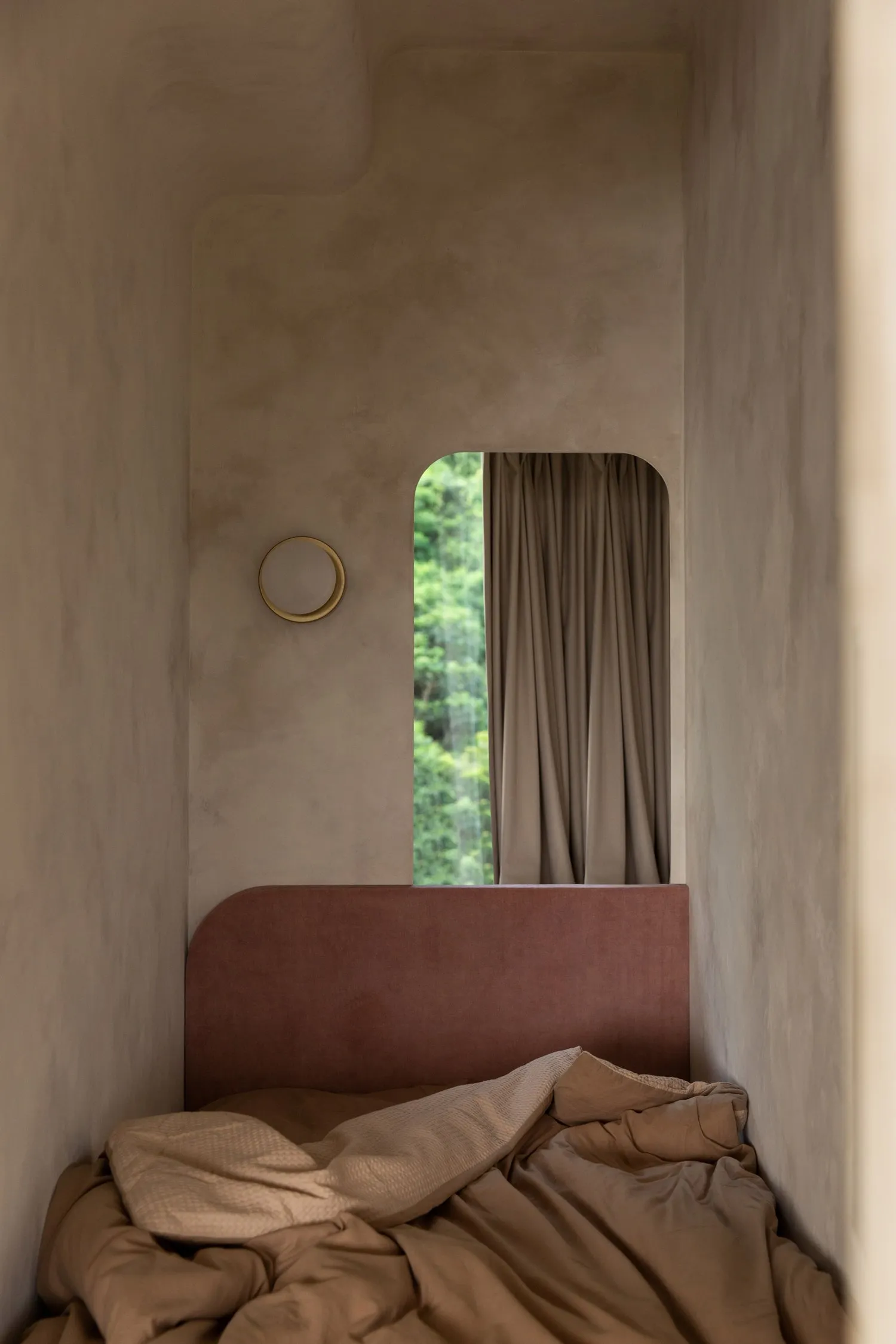 A minimalist bedroom with a pink velvet headboard and a cozy atmosphere, featuring a round light on the wall and a view of gr