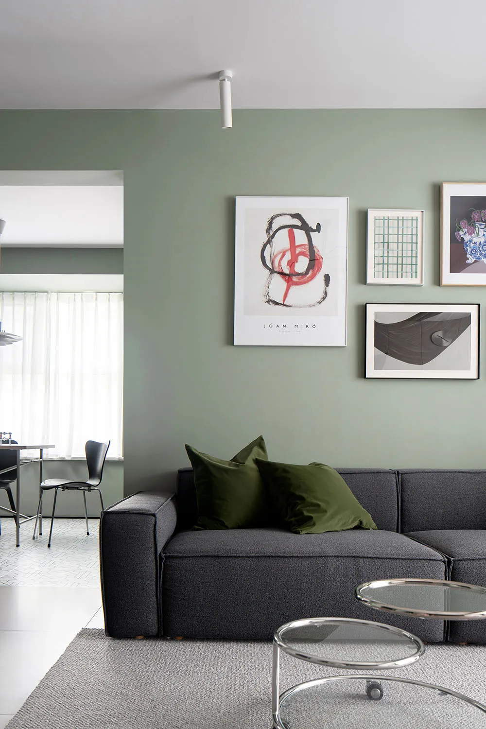 A modern living room featuring a grey sofa with green cushions, framed artworks on the wall, and glass coffee tables on a rug