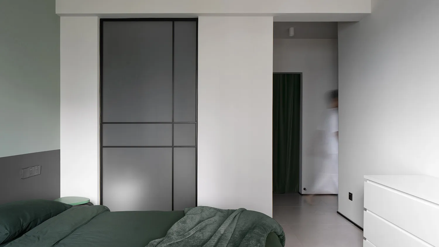 Minimalist bedroom with green bedding, black metal sliding doors, and white walls.