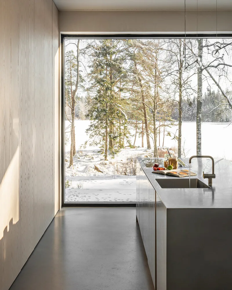 Modern kitchen with minimalist design featuring concrete countertops and floor, surrounded by a snowy forest view through lar