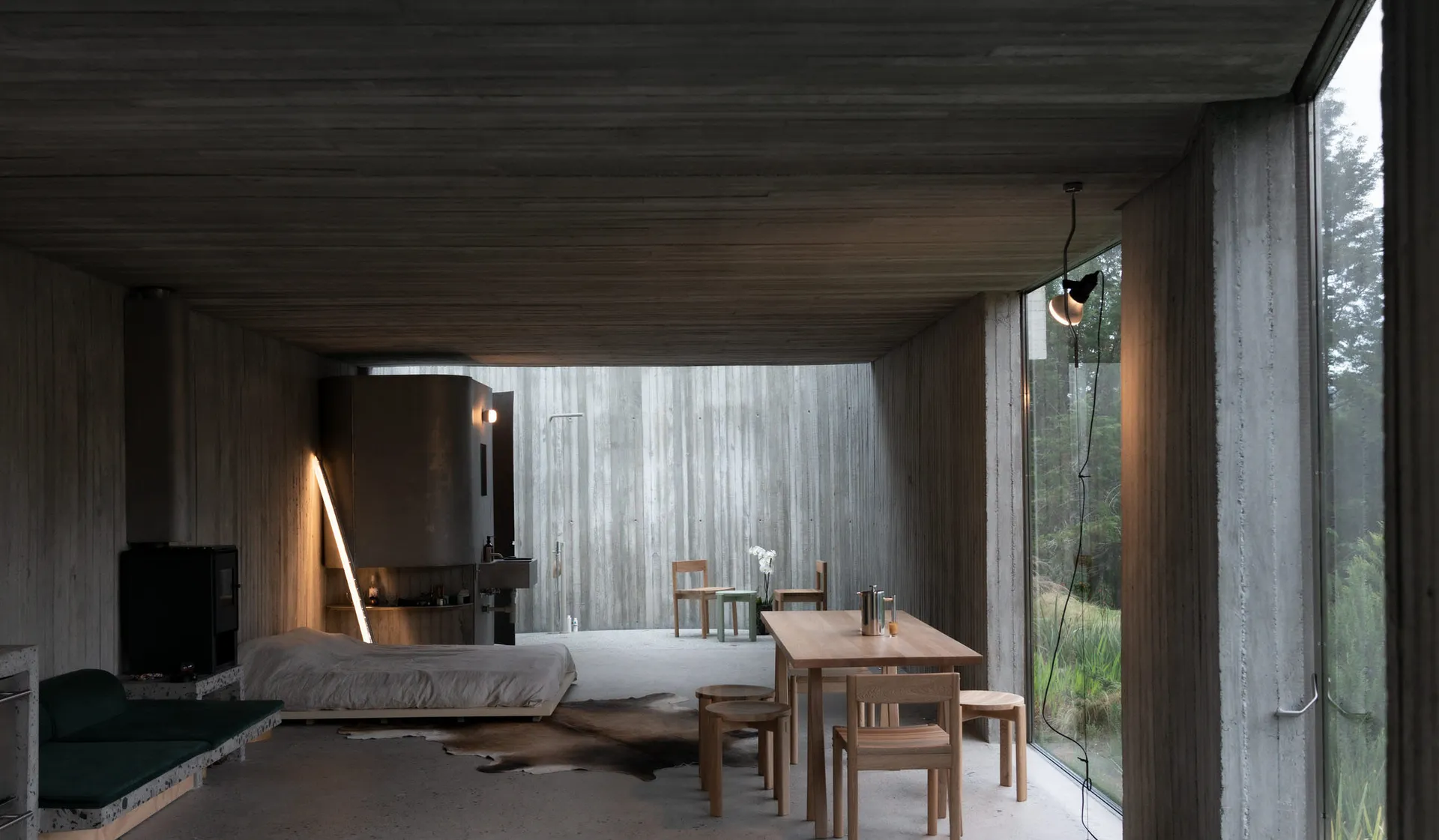A minimalist interior space with concrete walls and floor, featuring a table, chairs, and a bed.