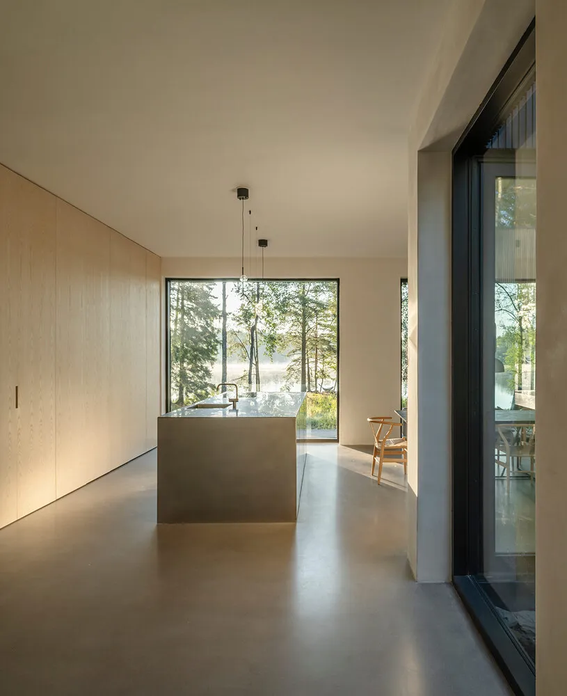 A modern residential kitchen featuring a concrete floor and island, with large windows showcasing a serene outdoor landscape.