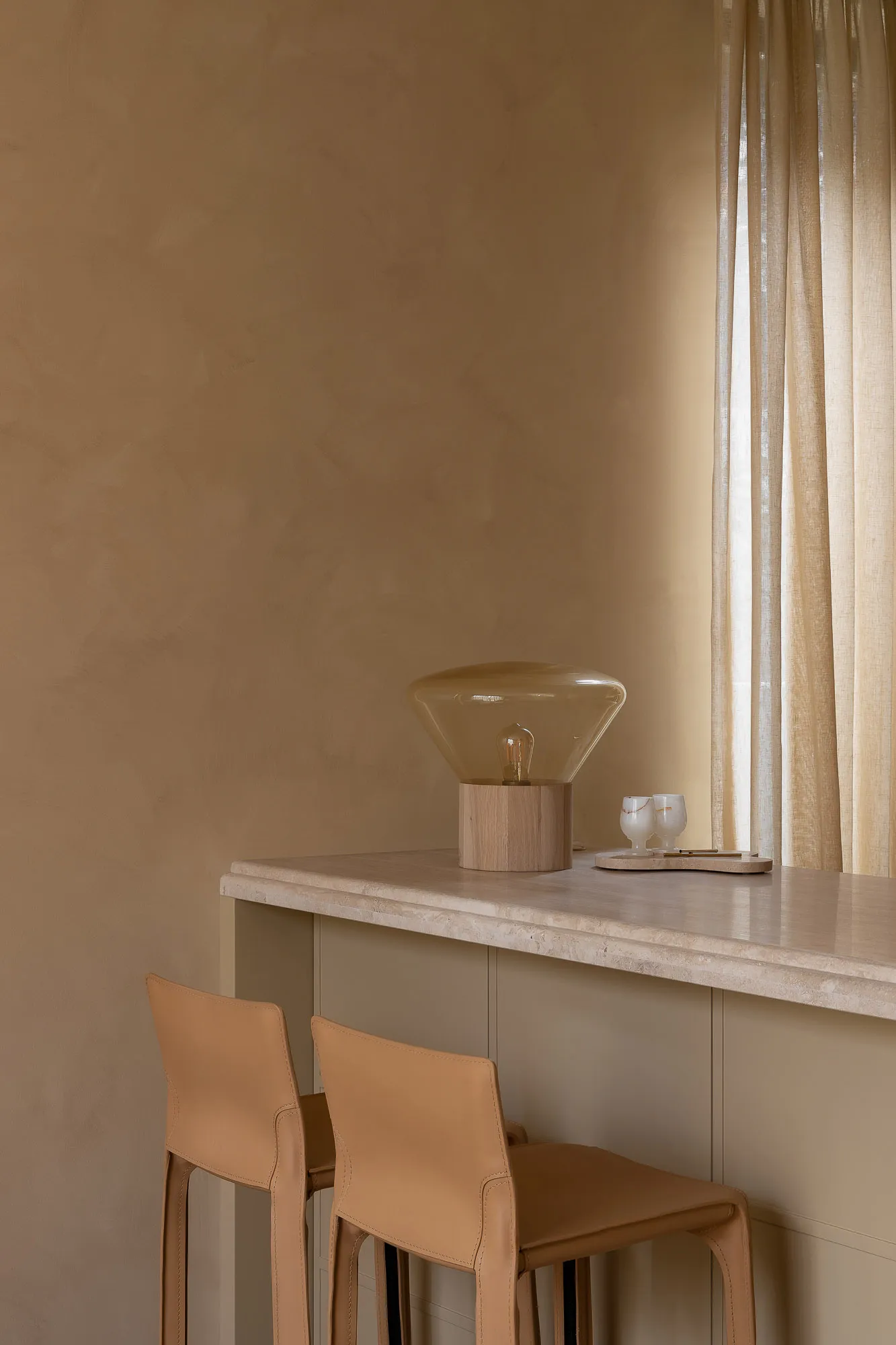 A minimalist kitchen featuring a concrete counter with a sleek, modern lamp and two white cups on a wooden tray.