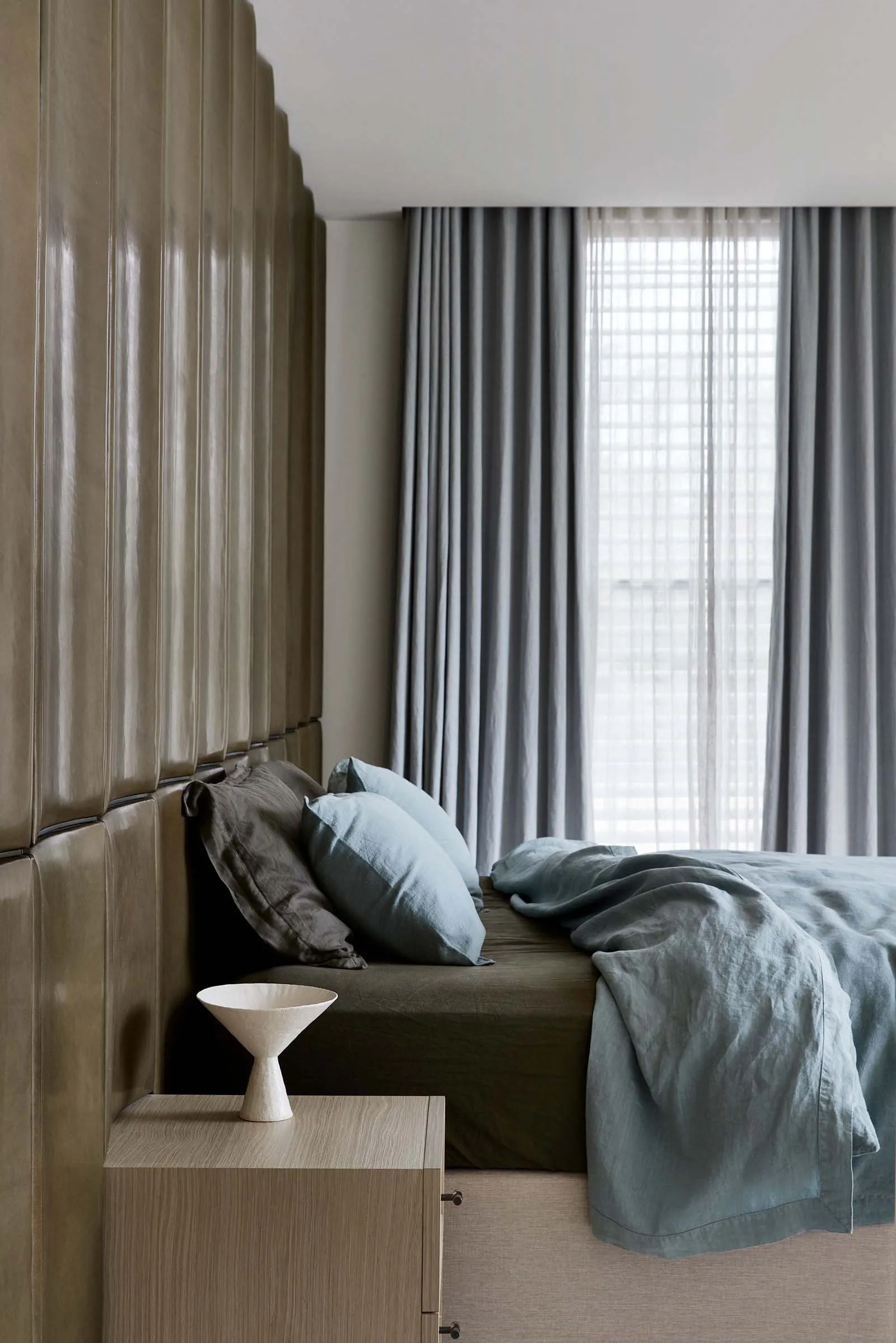 A modern minimalist bedroom with fabric headboard, light grey curtains, and light blue bedding.