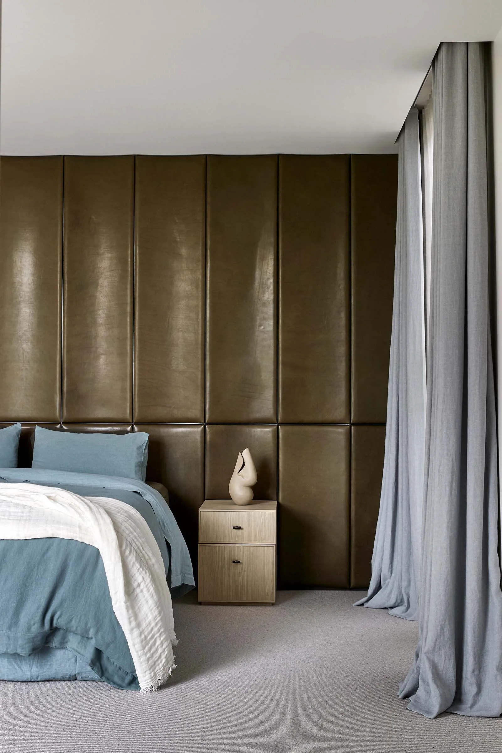 A modern minimalist bedroom featuring a large bed with blue bedding, a side table with a sculpture, and a leather accent wall