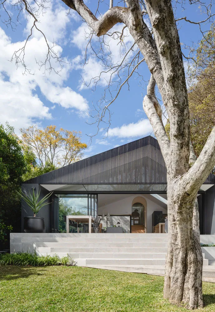 A modern house with a metal-clad exterior, featuring large glass windows and a sloping roof surrounded by greenery.