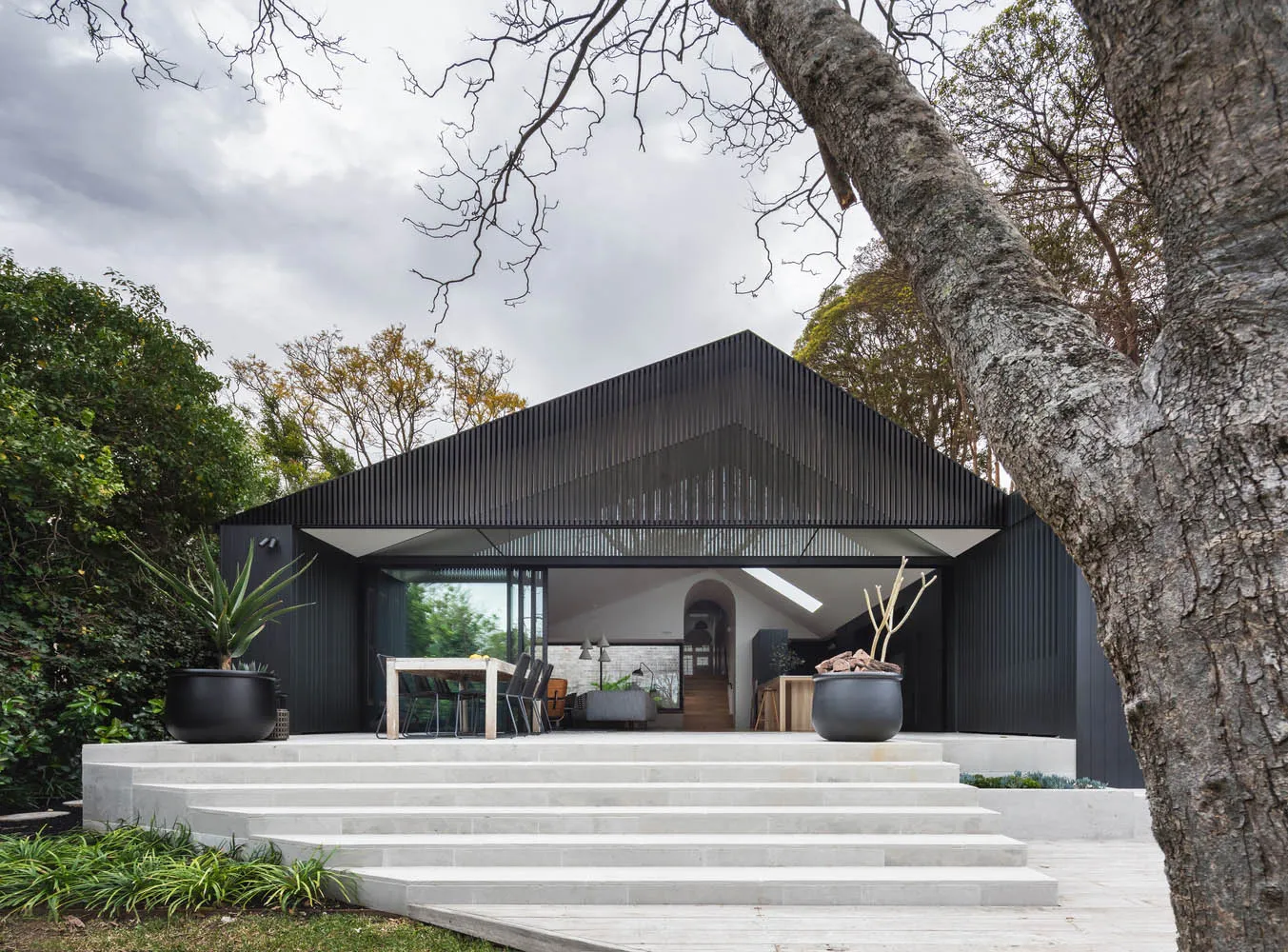 A modern residential exterior with black corrugated metal siding, large glass windows, and a large tree in the foreground.