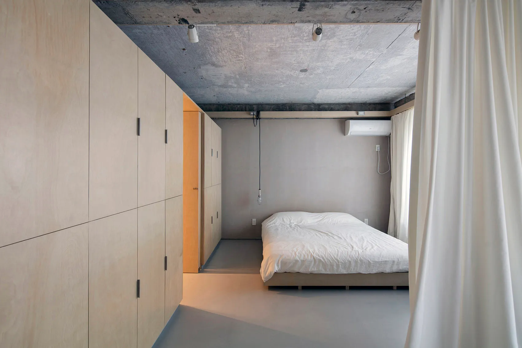 A minimalist bedroom with a concrete floor and walls, featuring a plain white bed with a simple wooden frame.