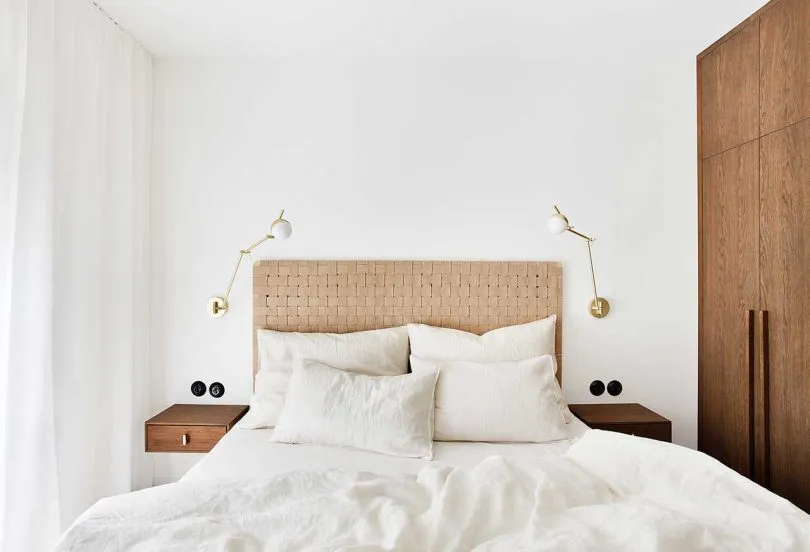 Minimalist bedroom featuring a woven headboard, white linen bedding, and wooden nightstands with wall-mounted lamps.