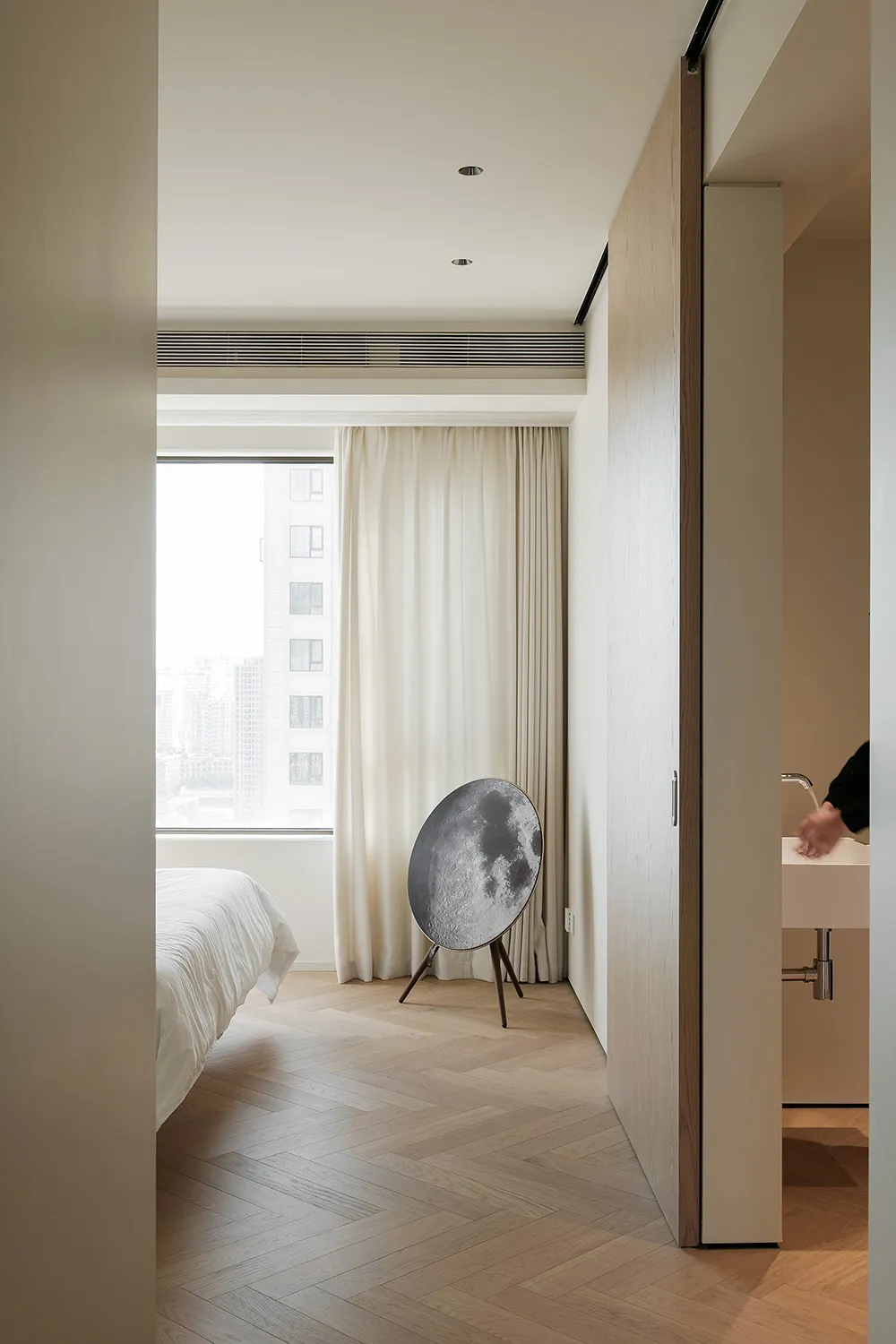 A modern minimalist bedroom with white walls, white curtains, a white bed, and a circular speaker with a moon pattern on a wo