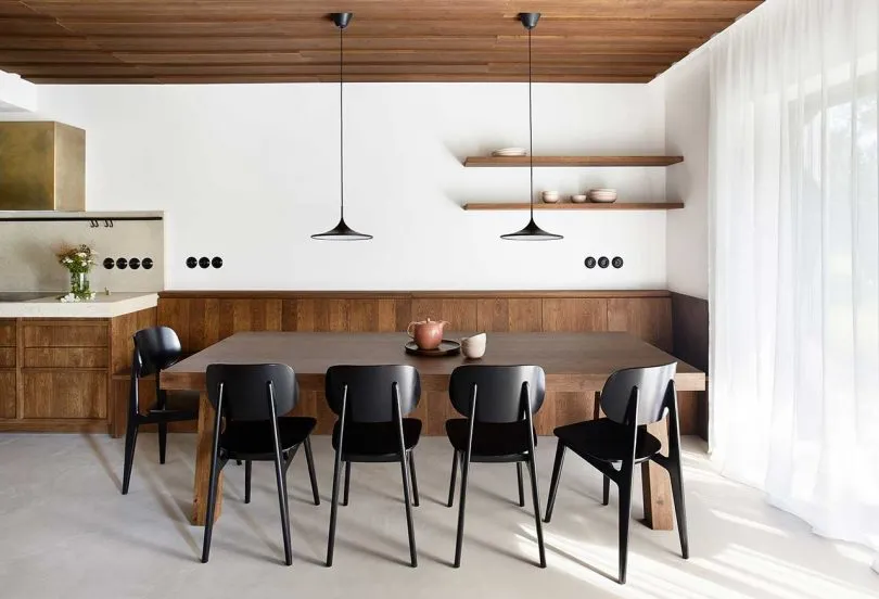 A modern dining room featuring a wooden table and chairs, with a white curtain and floating shelves.
