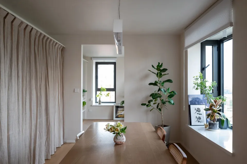 Modern living room featuring a wooden dining table adorned with flowers, surrounded by plants and glass window, with a fabric