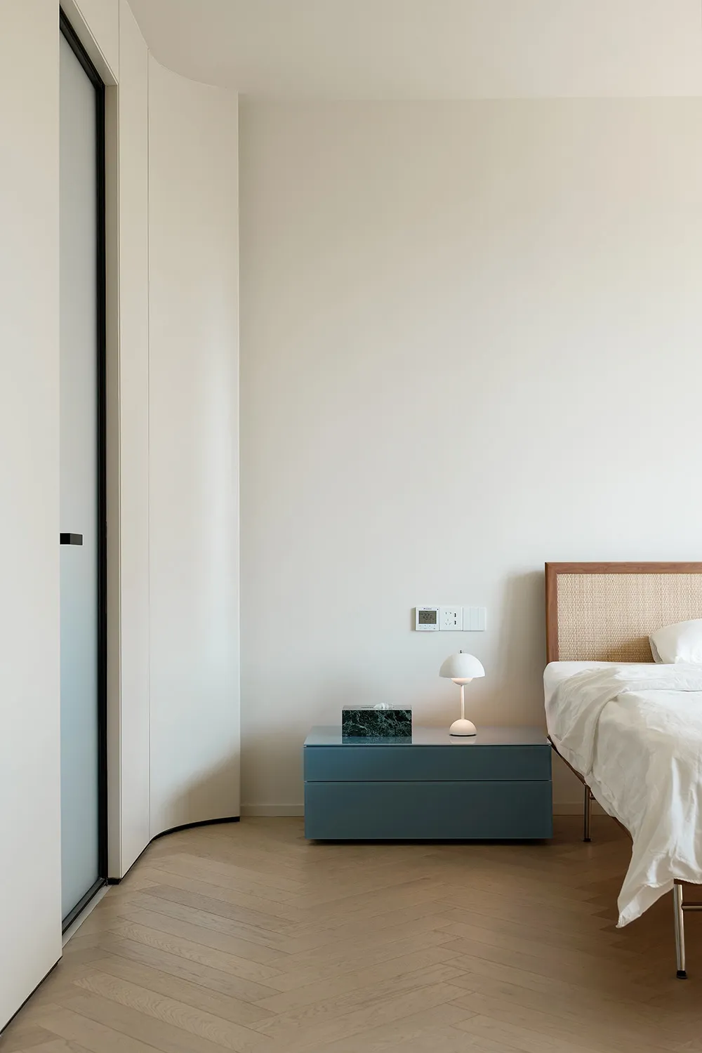 A modern, minimalist bedroom featuring a bed with a white fabric headboard and a nightstand with a blue drawer and a white la