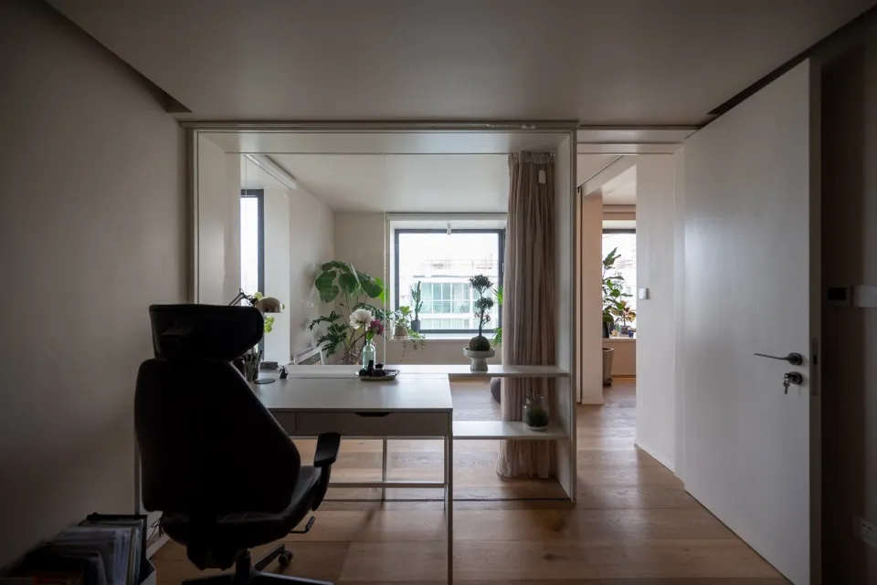 A minimalist home office with a white desk and chair, surrounded by plants and natural light.