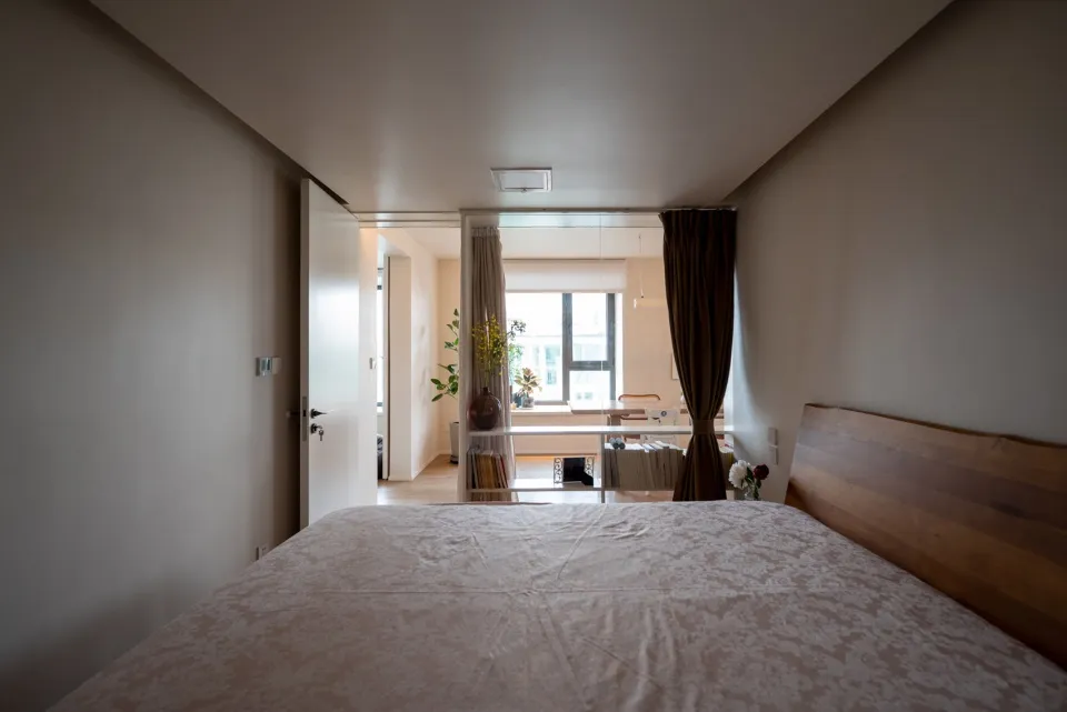 A minimalist bedroom with a wooden headboard, beige fabric bedding, and subtle patterned bedding spread across a bed, with a