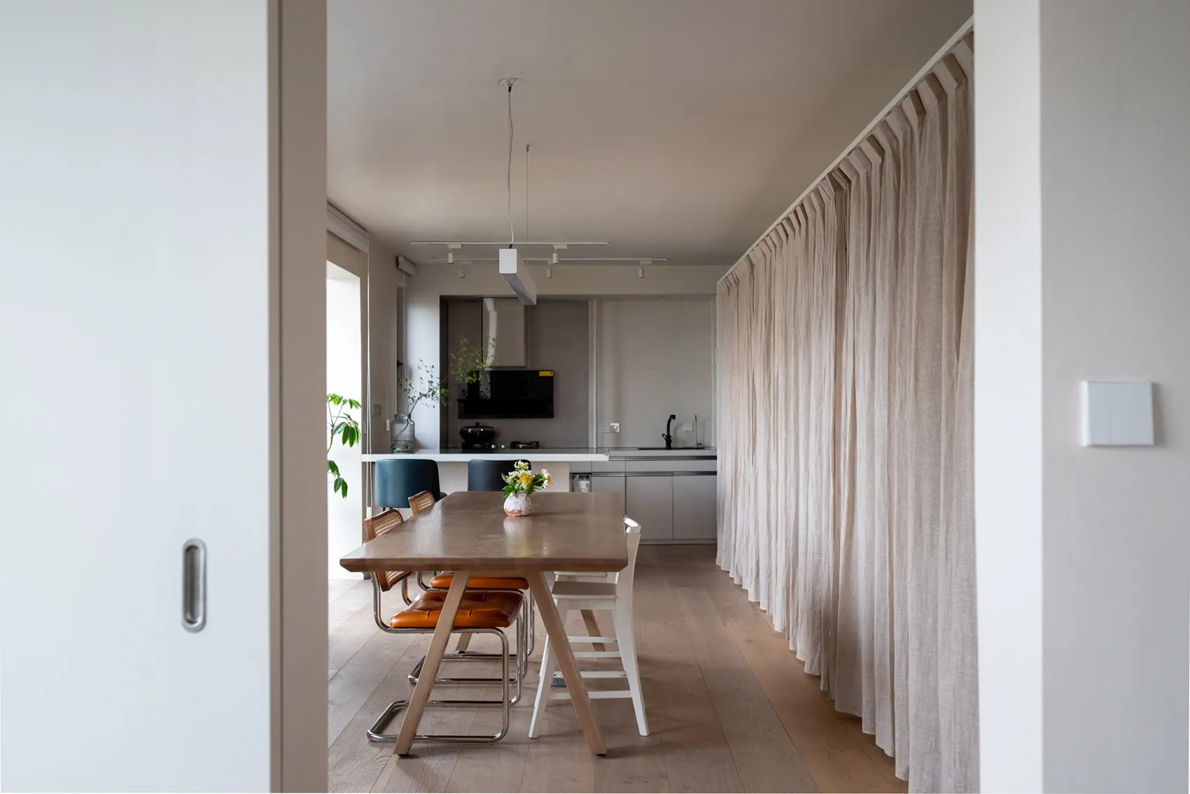 A modern minimalist kitchen with a wooden table, fabric curtains, and metal chairs.