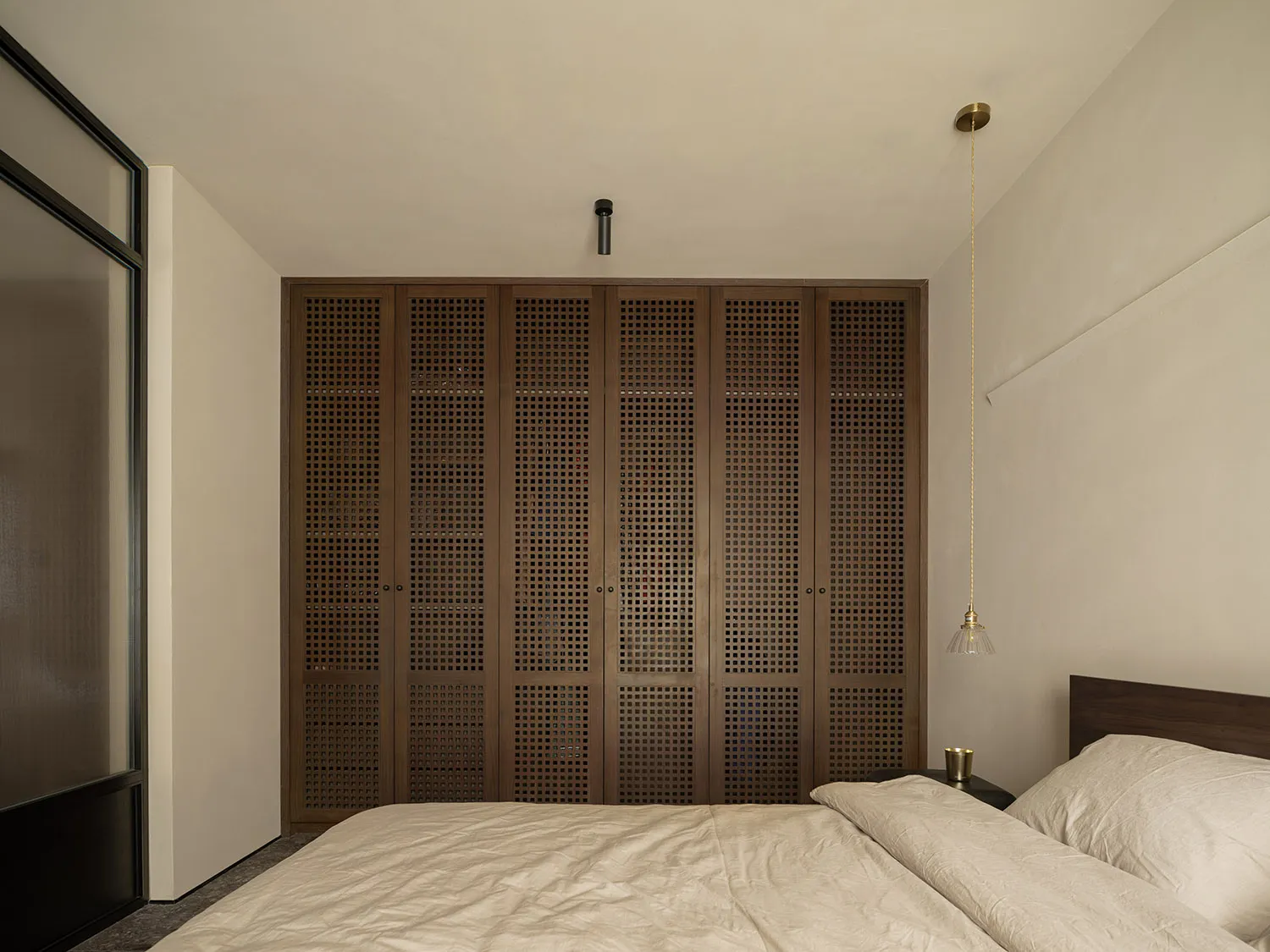 A minimalist bedroom featuring wooden paneled storage cabinets, a bed with a beige comforter, and a hanging lamp.