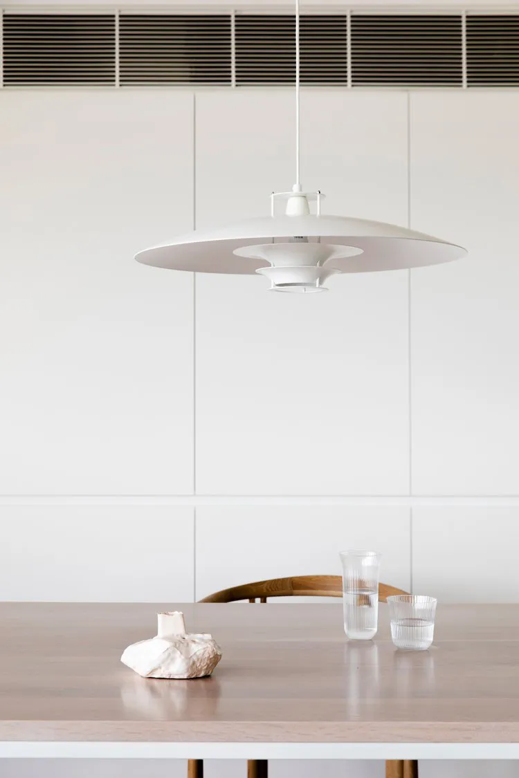 A minimalist interior space featuring a white table with a wooden texture, a white pendant light fixture, and glass cups.