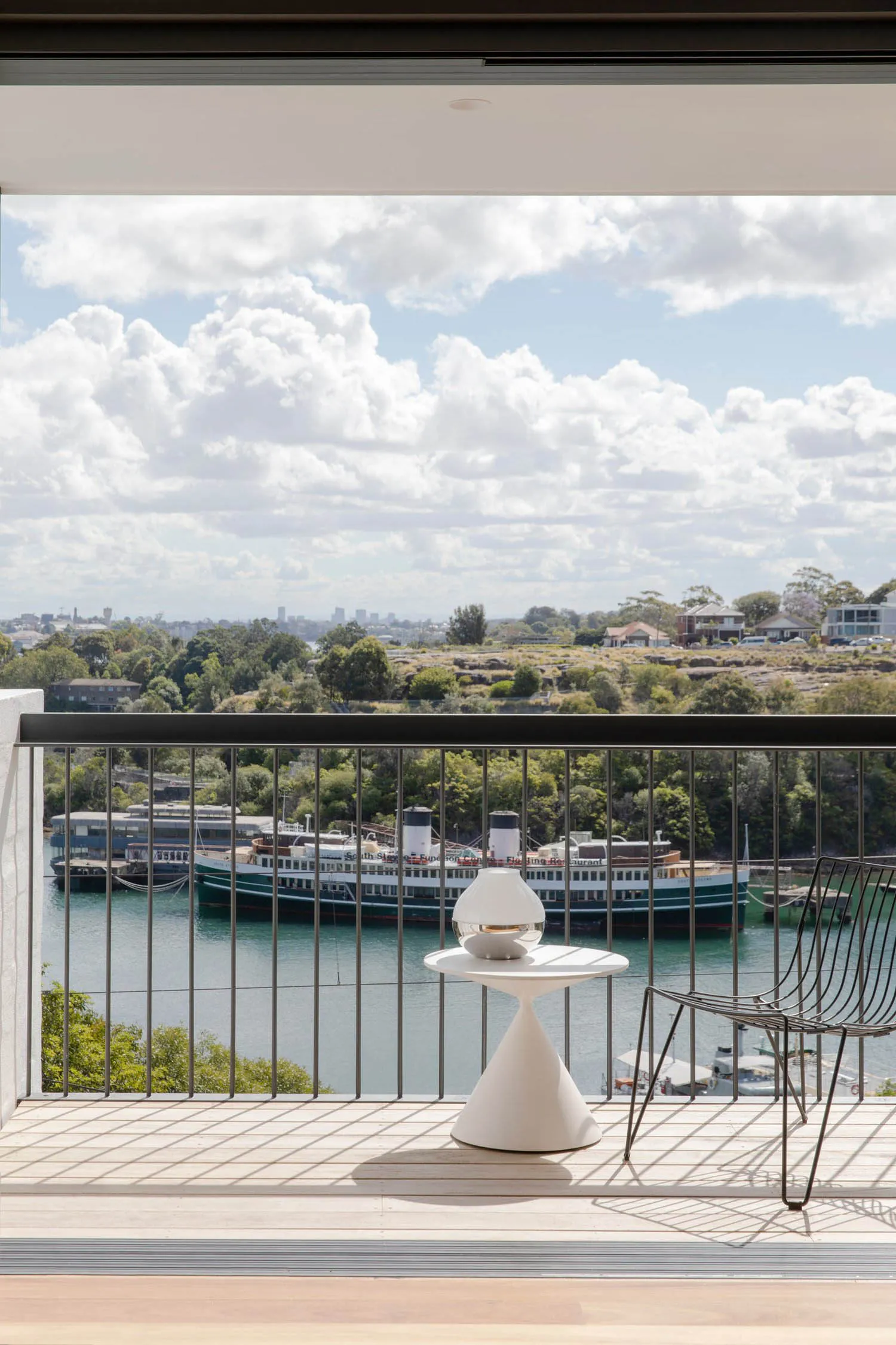 A modern balcony featuring a metal table and chair, with a scenic river view and ships in the background.