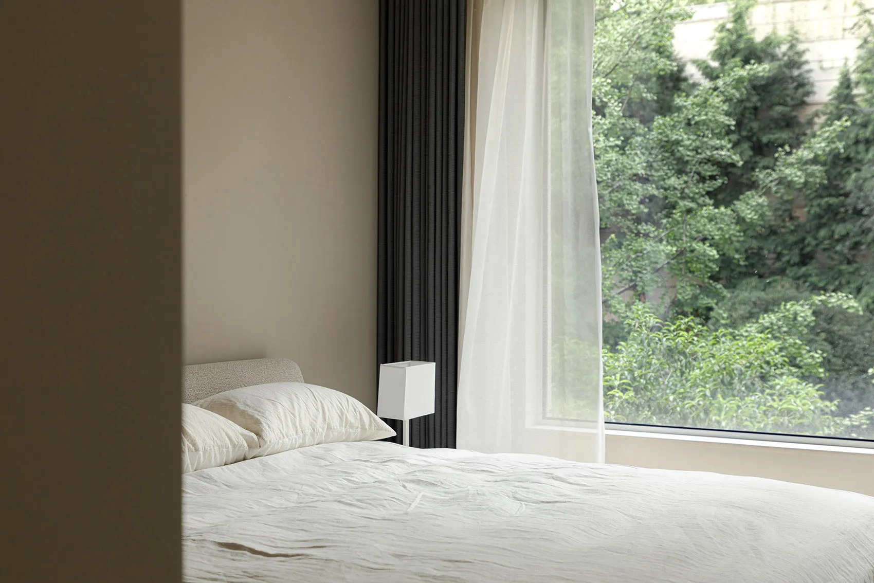 A minimalist bedroom with a bed featuring white textiles and natural light streaming through a large window with a sheer curt