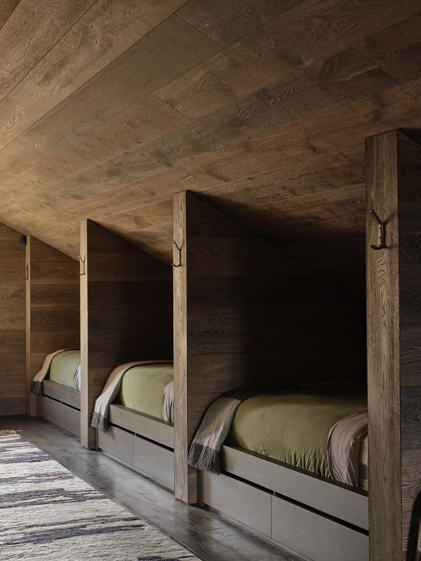 A minimalist bedroom featuring wooden walls and floors, with three single beds in a slanted room.