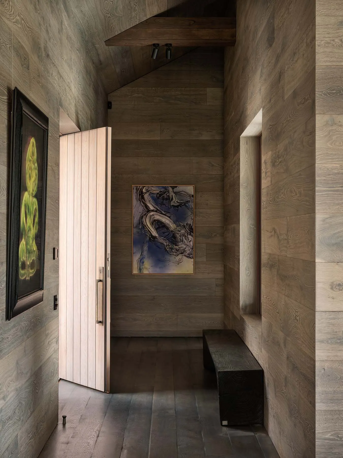 A modern entryway featuring wooden walls and a door, with abstract artwork and a bench.