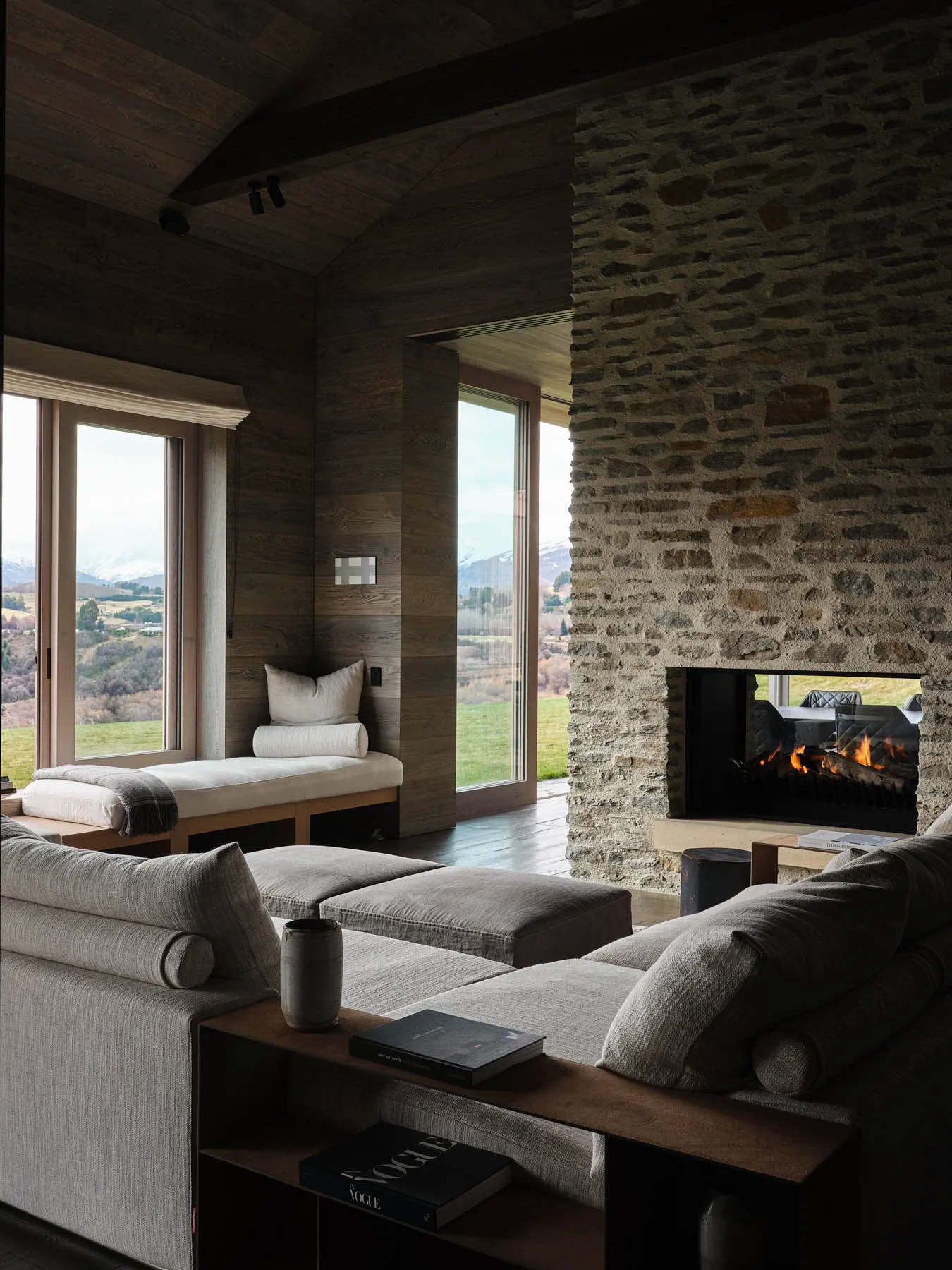 Modern living room featuring a stone fireplace and wood-paneled walls, with a view of the landscape beyond the large windows.