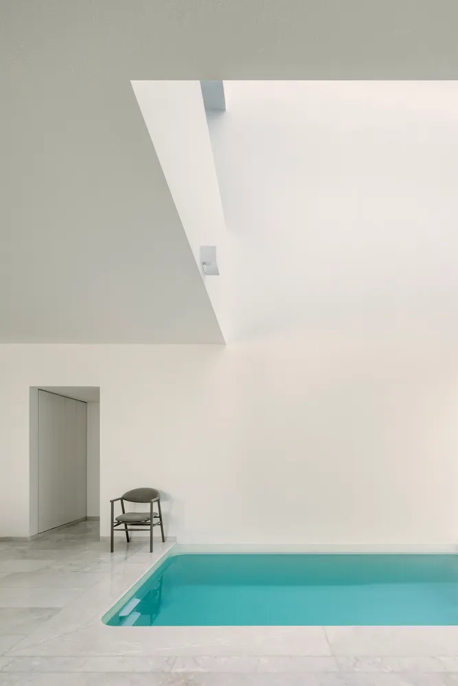A minimalist indoor swimming pool with a white marble floor, surrounded by white walls and a ceiling with a skylight.