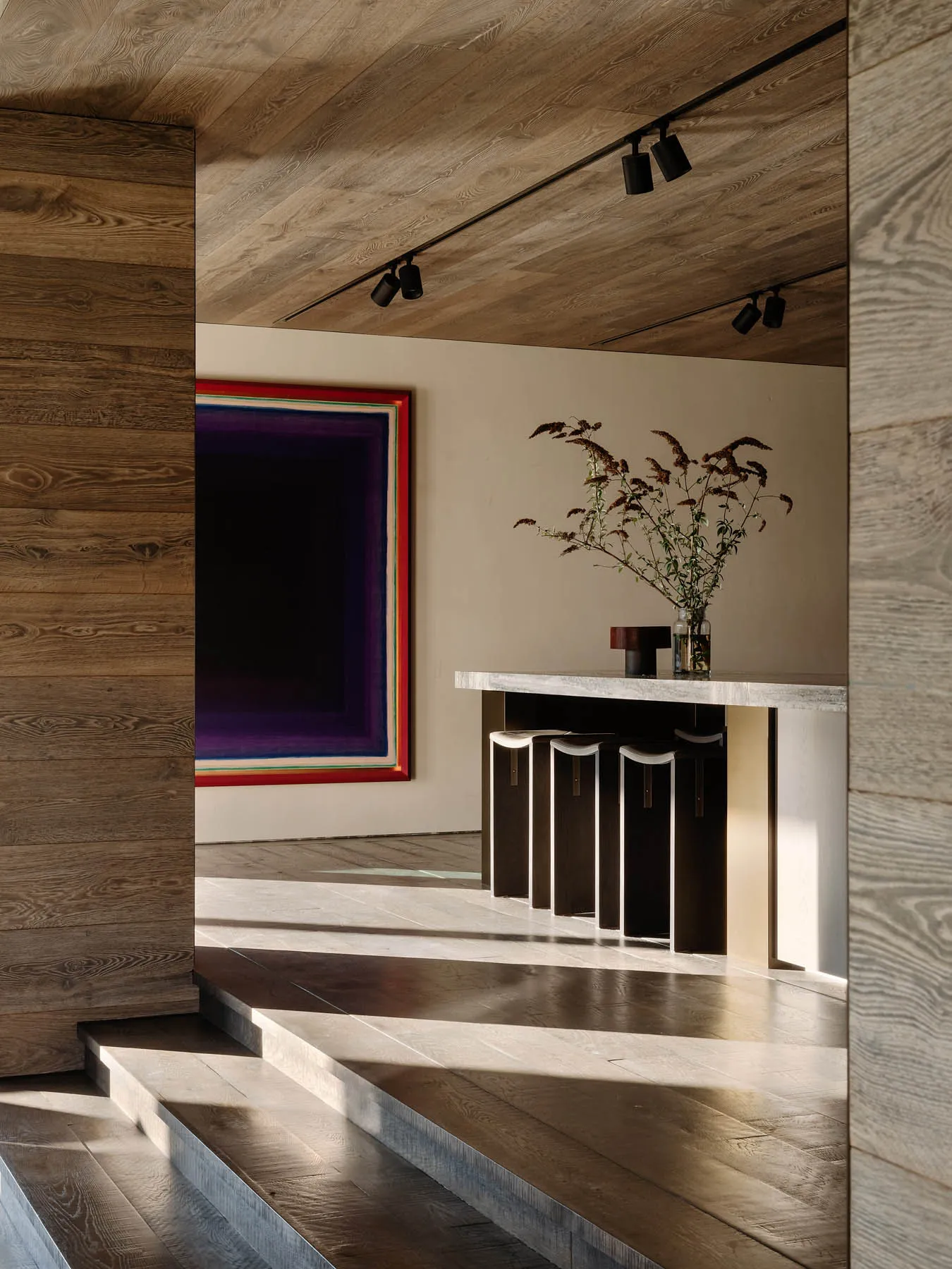 Modern residential interior with wooden flooring, a minimalist bar area with stools, and a large colorful artwork on the wall