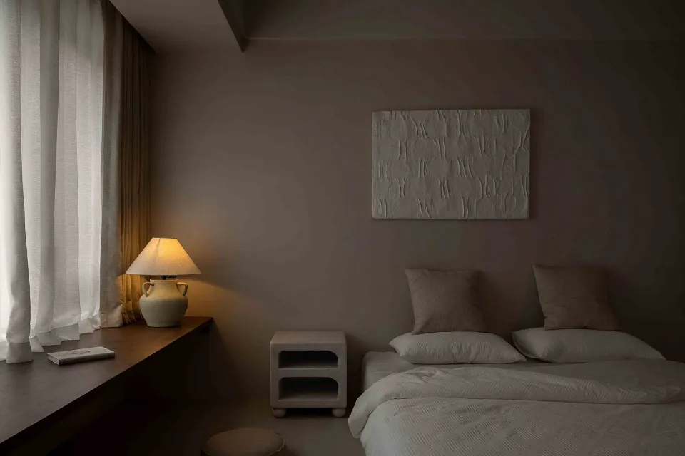 Minimalist bedroom with neutral tones, featuring a bed, nightstand, lamp, and artwork
