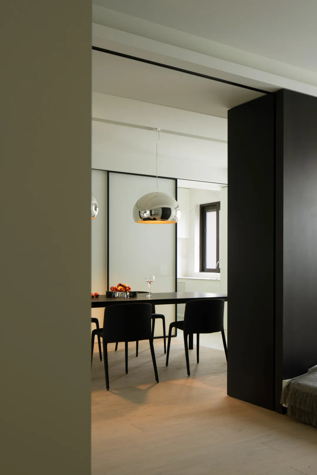 A modern minimalist dining room featuring a metallic pendant light fixture, black chairs, and a wooden floor.