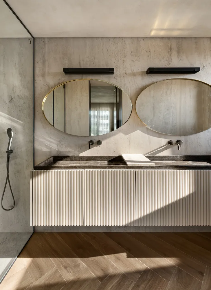 A minimalist bathroom featuring concrete walls, floating vanity with sleek white paneling, and oval mirrors above the sink. T