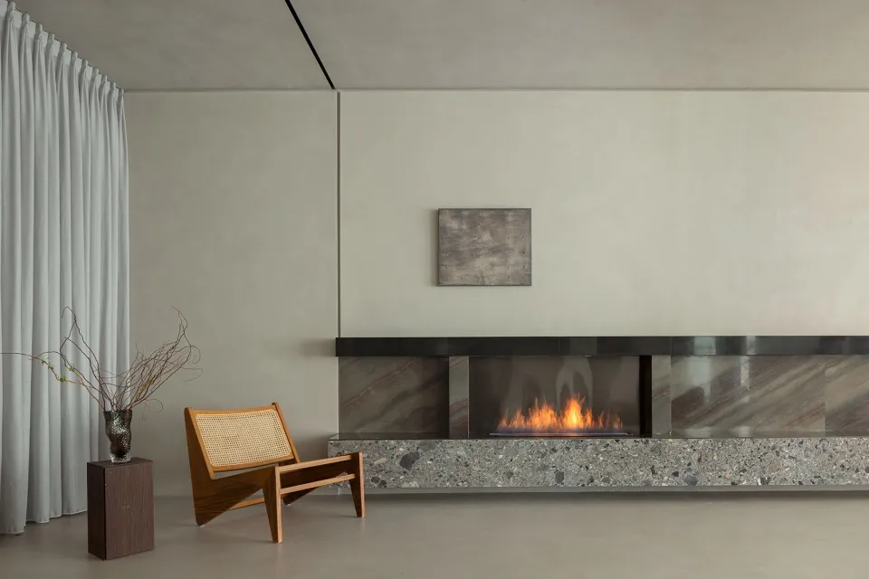 A modern living room featuring a marble fireplace with a glowing fire, a minimalist wooden chair, and gray curtains.