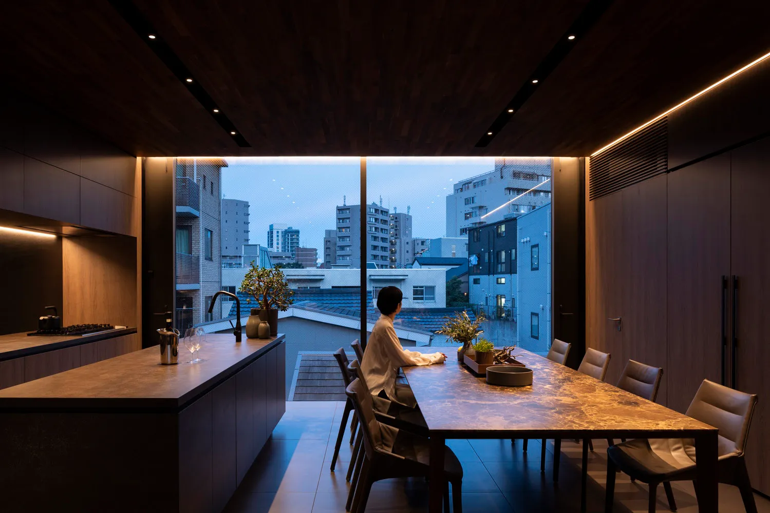 A minimalist kitchen with a marble countertop, modern seating, and a view of urban buildings through a large window.