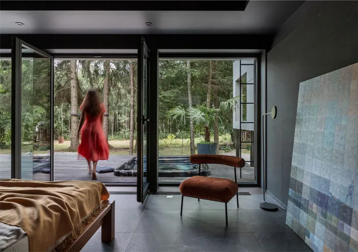A modern bedroom with concrete floors and a woman wearing a red dress walking on the wooden deck outside through large glass 