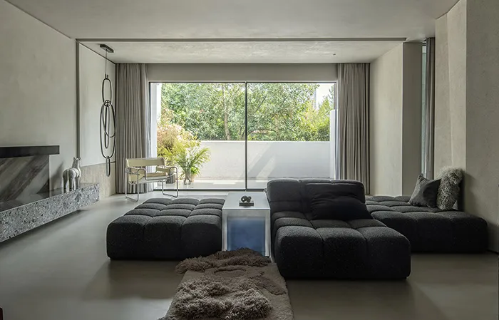 A modern minimalist living room with a concrete floor, large windows, and a large black modular sofa.