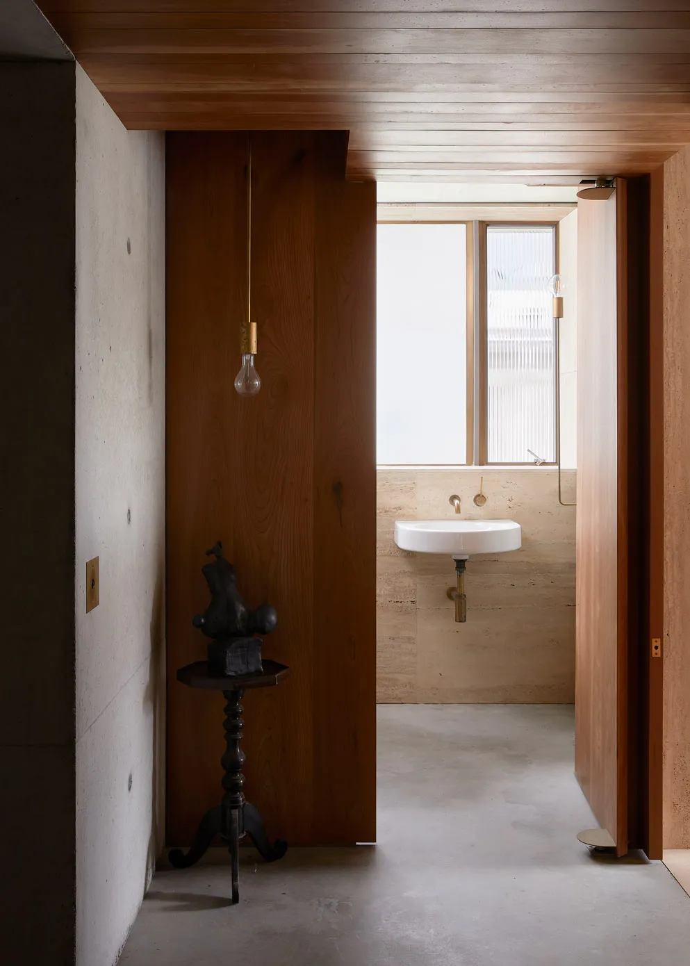 A modern minimalist bathroom with concrete walls and floor, featuring a wall-mounted sink and a hanging light bulb.