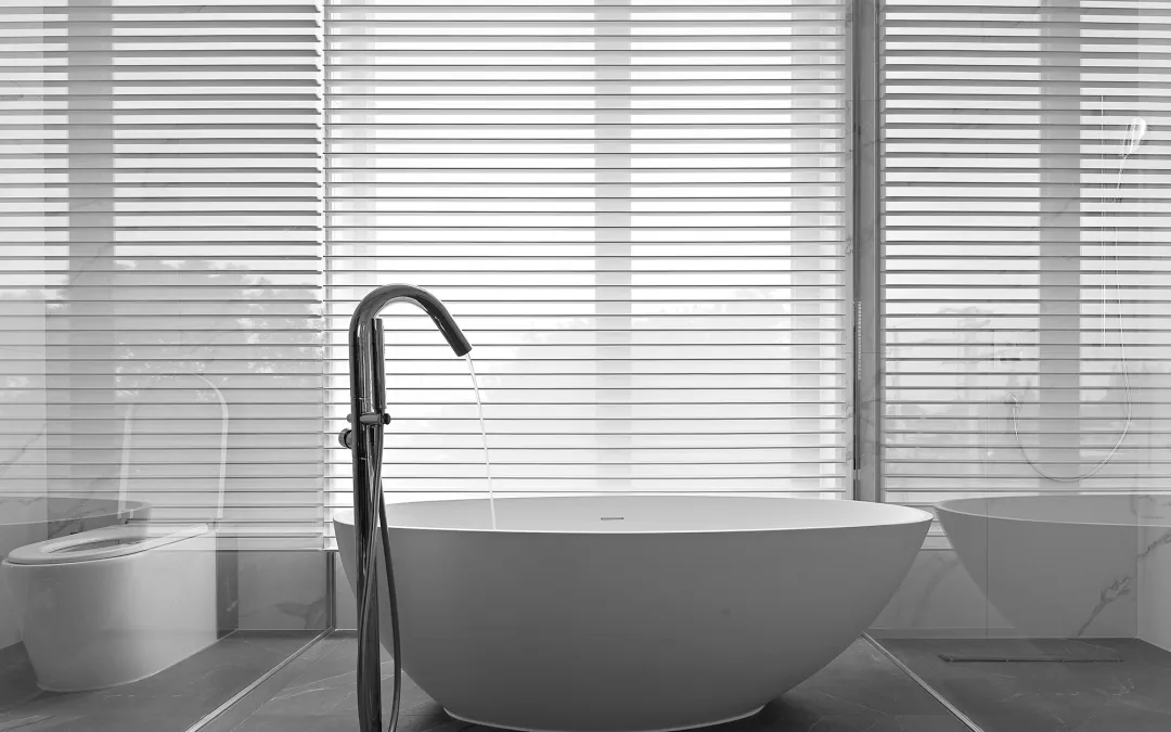 Modern bathroom with a white ceramic freestanding bathtub in front of a window with white blinds.