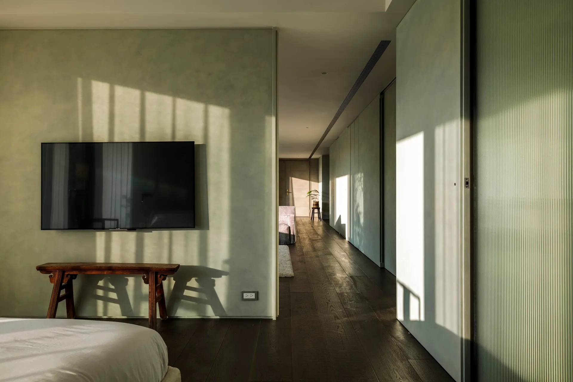 Modern, minimalist interior with concrete finishes, a white bed, and a wooden bench in front of a wall-mounted television.
