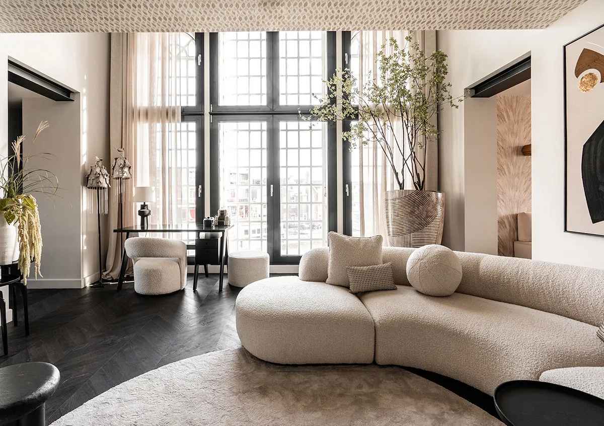 Modern living room featuring a curved, beige fabric sofa, dark wood flooring, and large windows with white curtains.