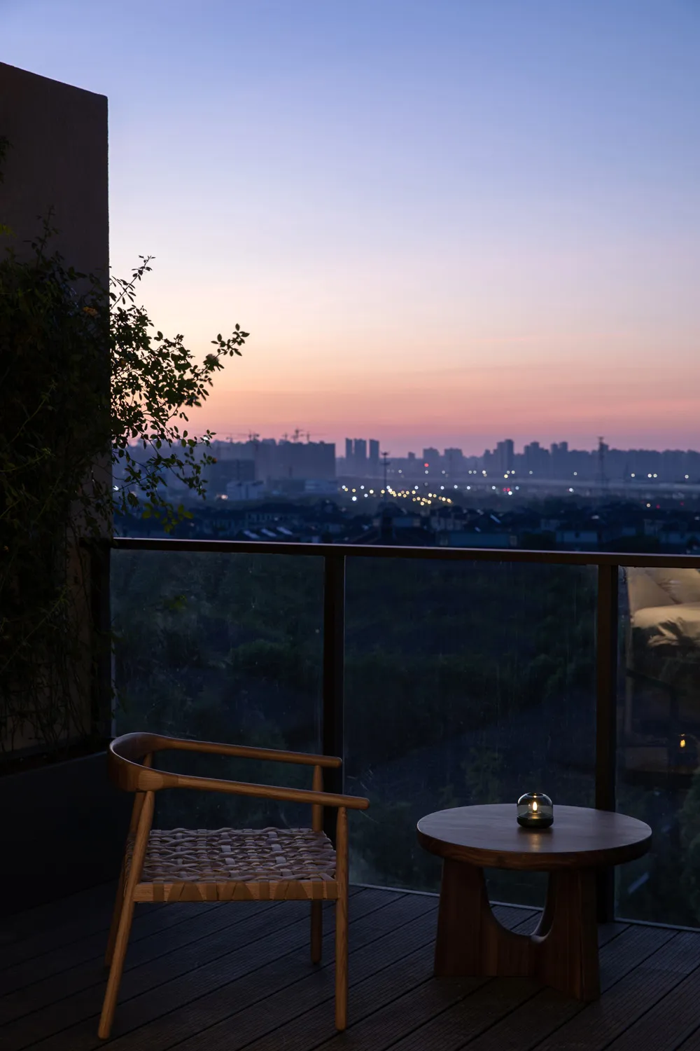 A wooden chair with a woven seat sits on a wooden deck overlooking a cityscape at dusk, illuminated by ambient light.