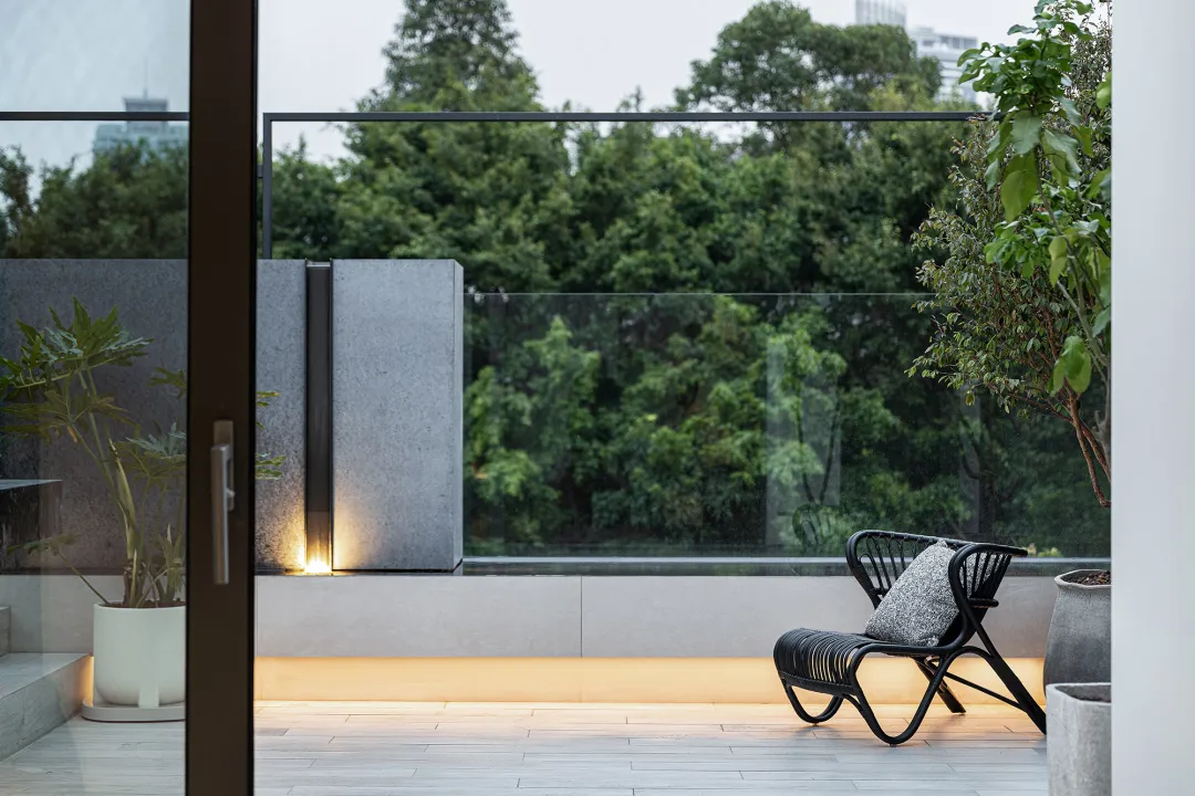 Black wicker chair with white cushion on a concrete patio bordered by glass railings, surrounded by lush greenery and trees.