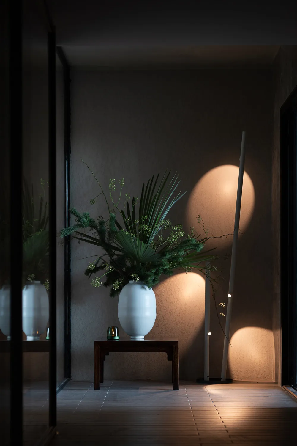 A wooden table with a white vase holding green plants, a lamp with a cylindrical design casting light on the wall, and a wood