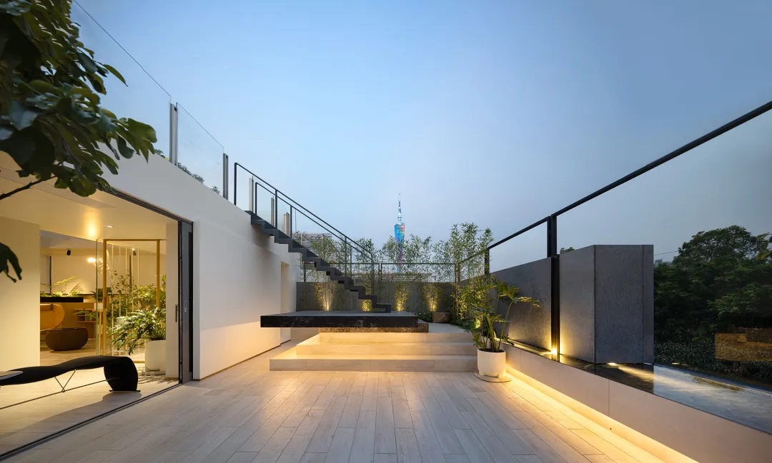 A modern rooftop garden extension with wooden floors, concrete walls, and glass railings illuminated by warm lighting.