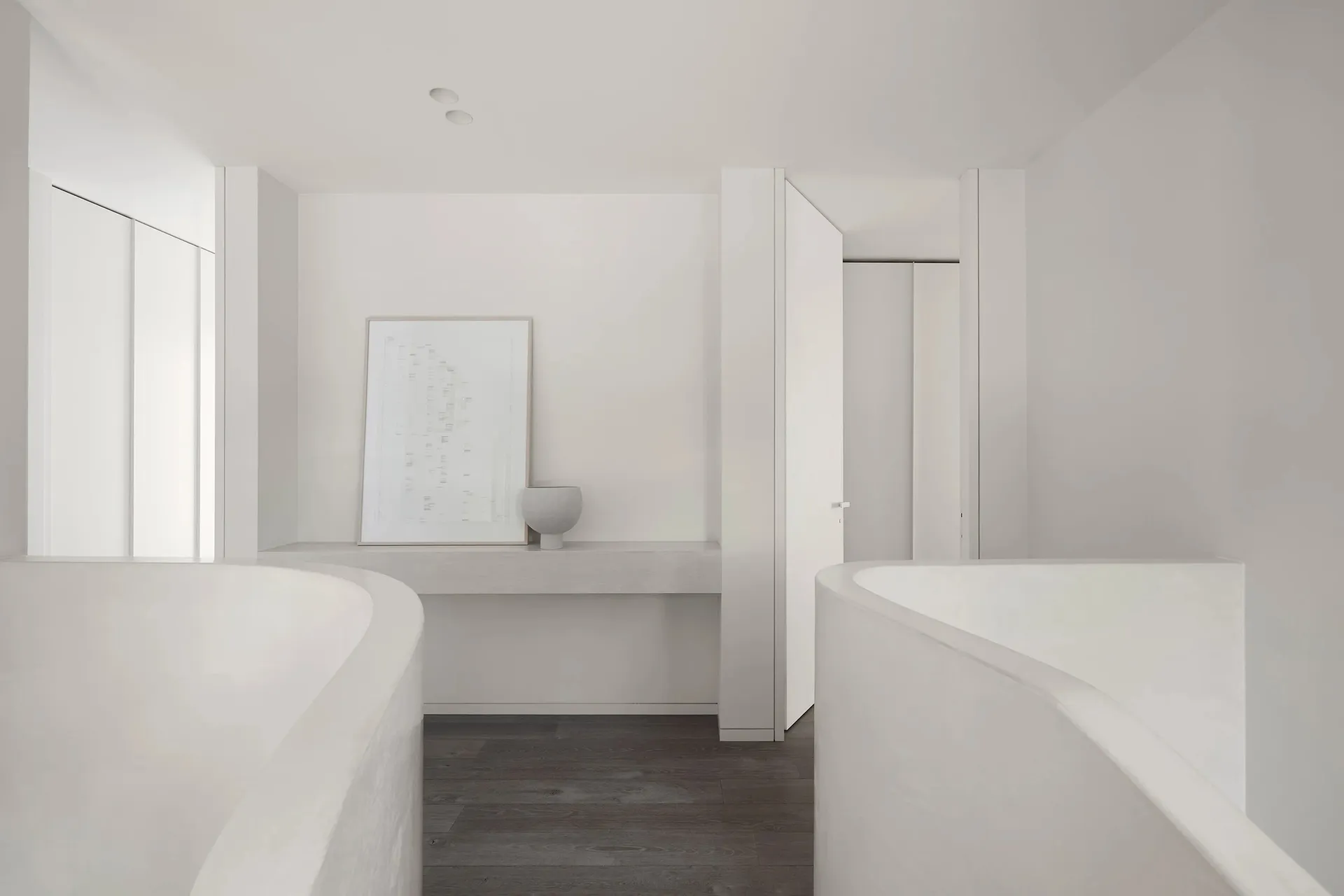 A minimalist bathroom with two concrete bathtubs, dark wooden flooring, and a small shelf with a vase and framed artwork.