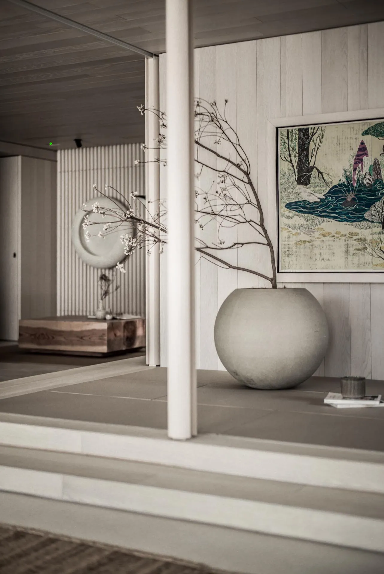 A minimalist interior design featuring a concrete vase with a bare branch and a framed painting hanging on the wall.