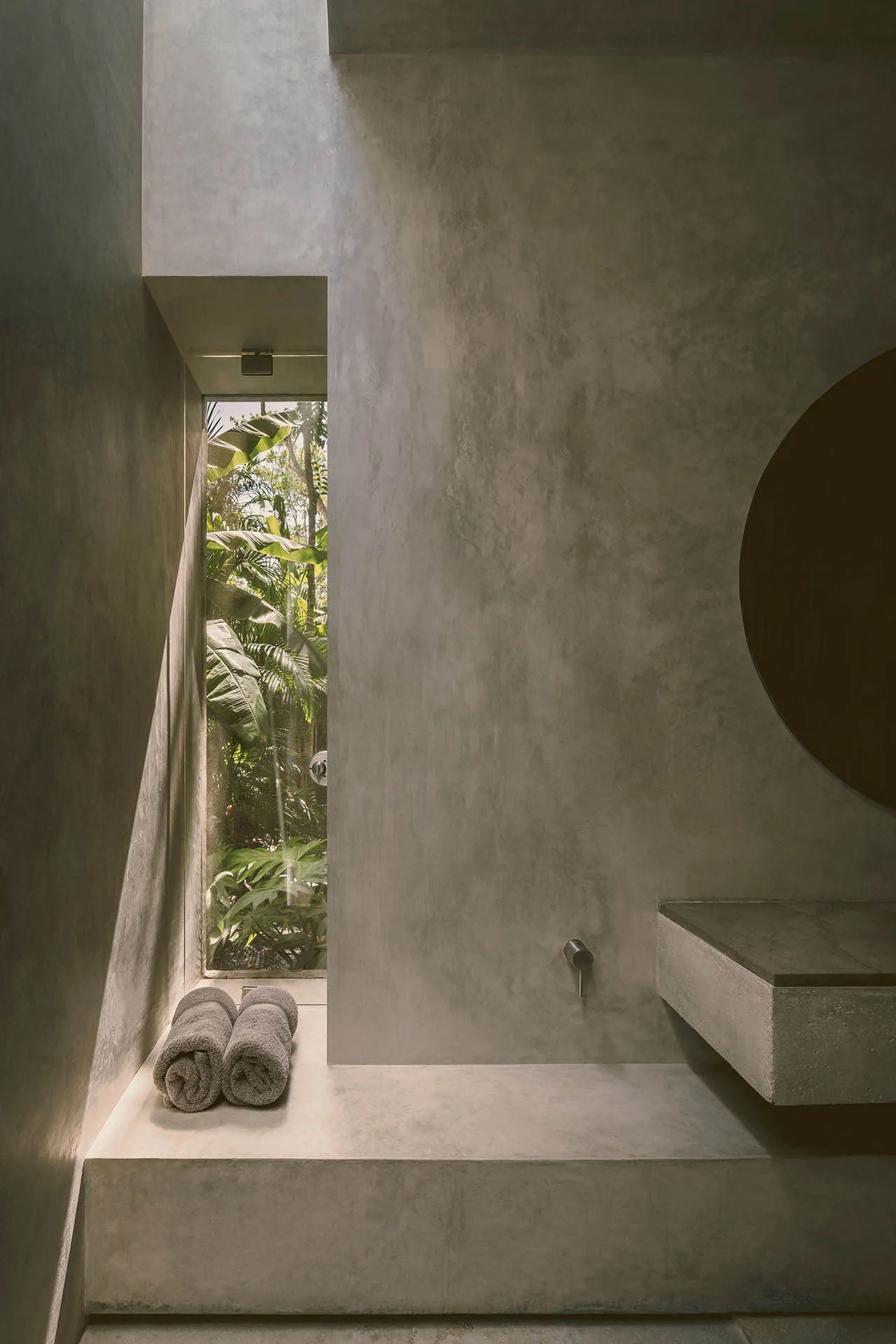 A modern bathroom with concrete walls and a shower area that leads outside to lush greenery.